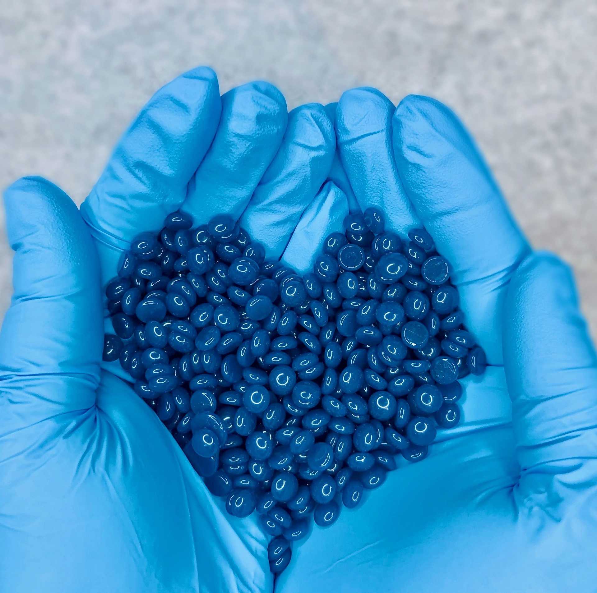 Hands with blue gloves holding a heart shape of blue plastic pellets against a gray background.