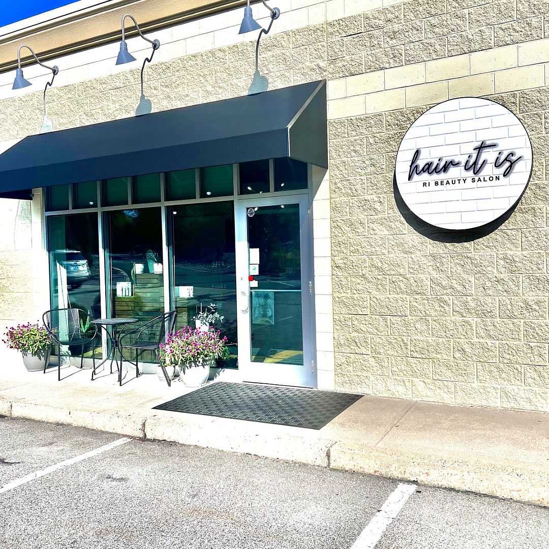 Front of Hair It Is beauty salon with black awning, round sign, and outdoor seating.