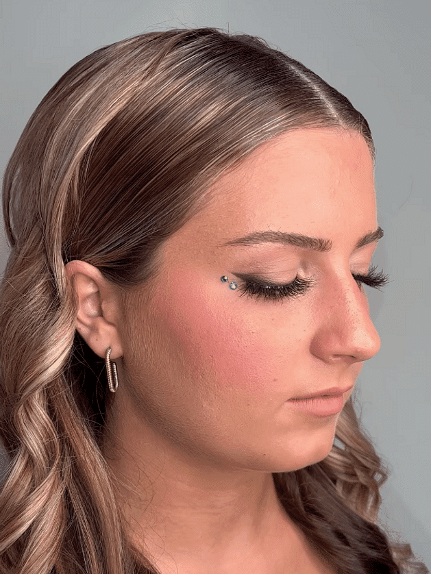 Woman with long hair, eyelash extensions, and facial gem, displayed in side profile.